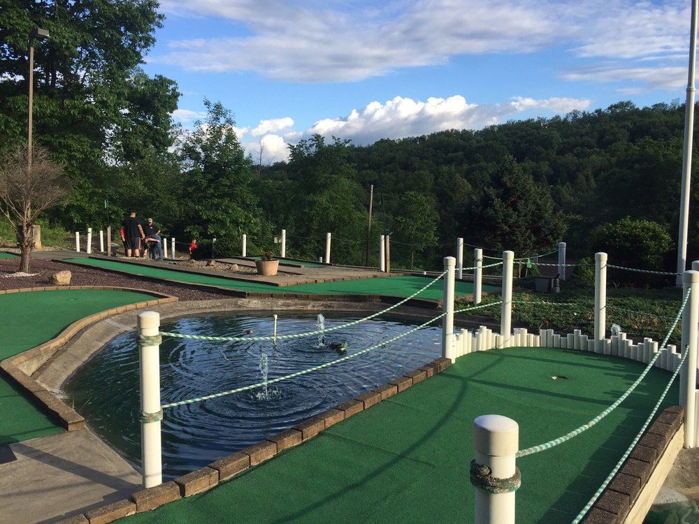 Mini golf course with water fountain feature surrounded by green carpet and trees